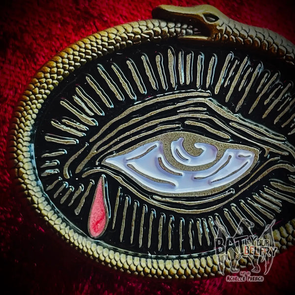 an antiqued gold metal pin of a weeping eye (illustrated in woodcut style) with a single iridescent red tear surrounded by a frame of a 3D sculpted ouroboros snake. Shown in close up