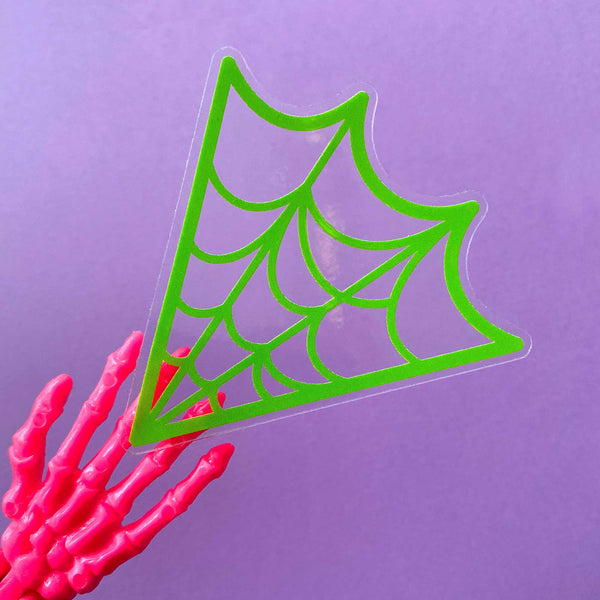 A die cut sticker of a neon green spiderweb on a clear background. Shown being held by a skeleton hand to show clear background against a purple backdrop 