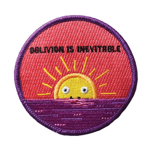 A round embroidered patch with purple border and a smiling yellow and orange sun rising from a purple horizon. The words “Oblivion is inevitable” are written in black above the sun.