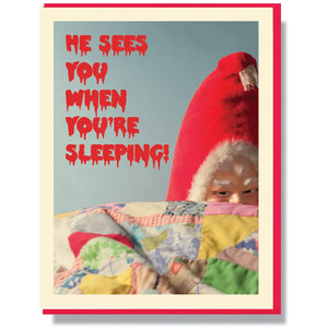 A rectangular holiday greeting card with a creepy Santa Claus doll peeking out from a quilt with the message “He sees you when you’re sleeping!” written in a red dripping blood style horror movie font