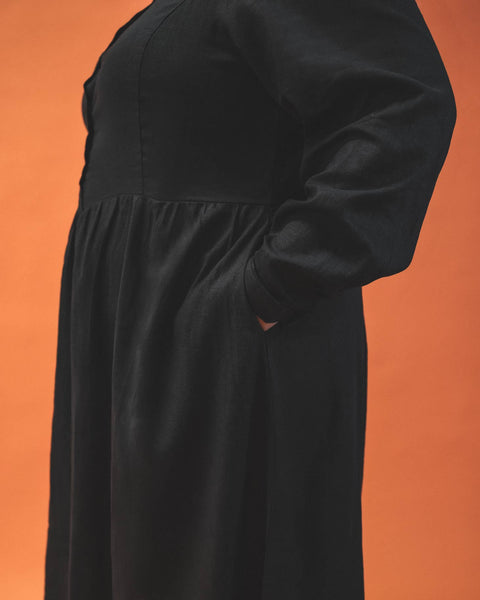 A plus size model wearing a long sleeved tea length linen shirtwaist dress with slightly puffed shoulders and princess seaming on the bodice. It has small matte black buttons down the front. The full skirt is slightly gathered. The model has their hands in both pockets and is seen zoomed in from the side