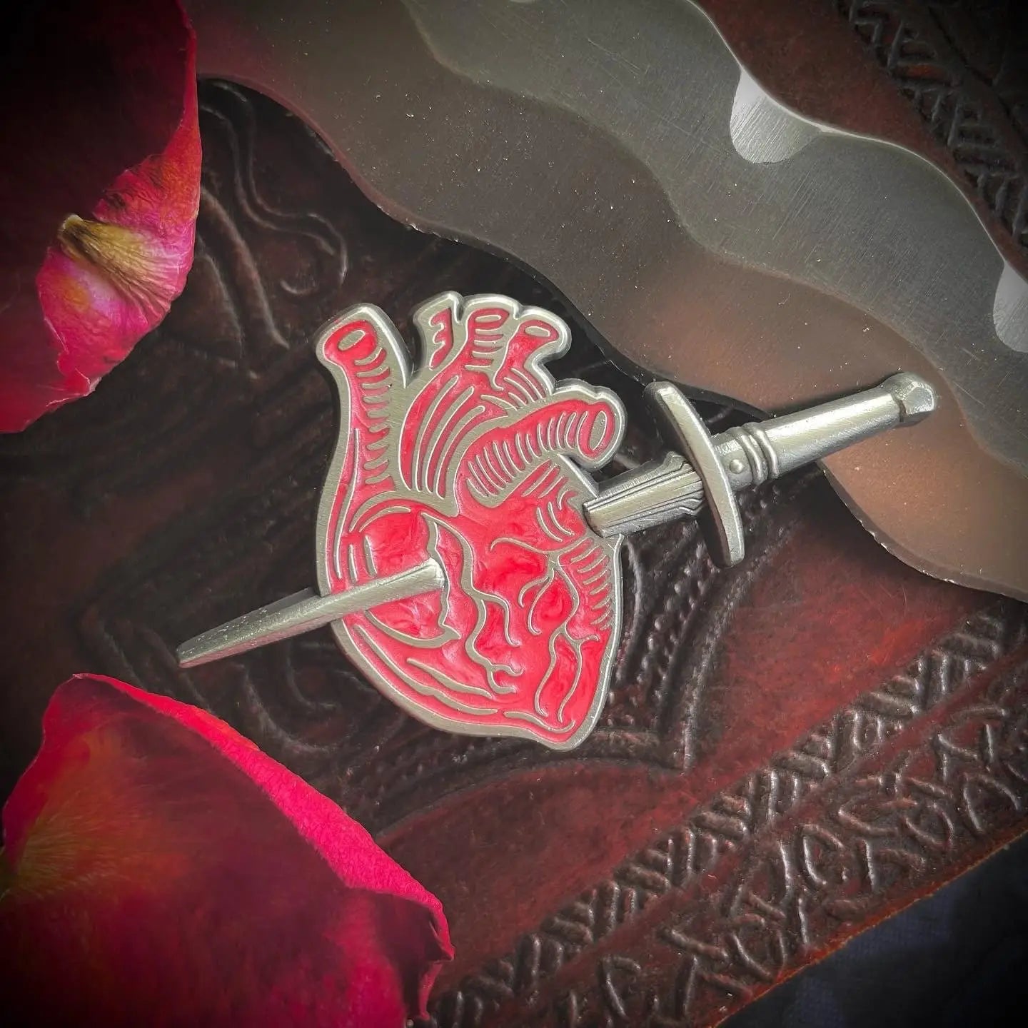 A pearly silver metal hard enamel pin with red detailing of an anatomical style heart being pierced by a 3D sculpted pointy dagger. Shown on a wooden table surrounded by rose petals