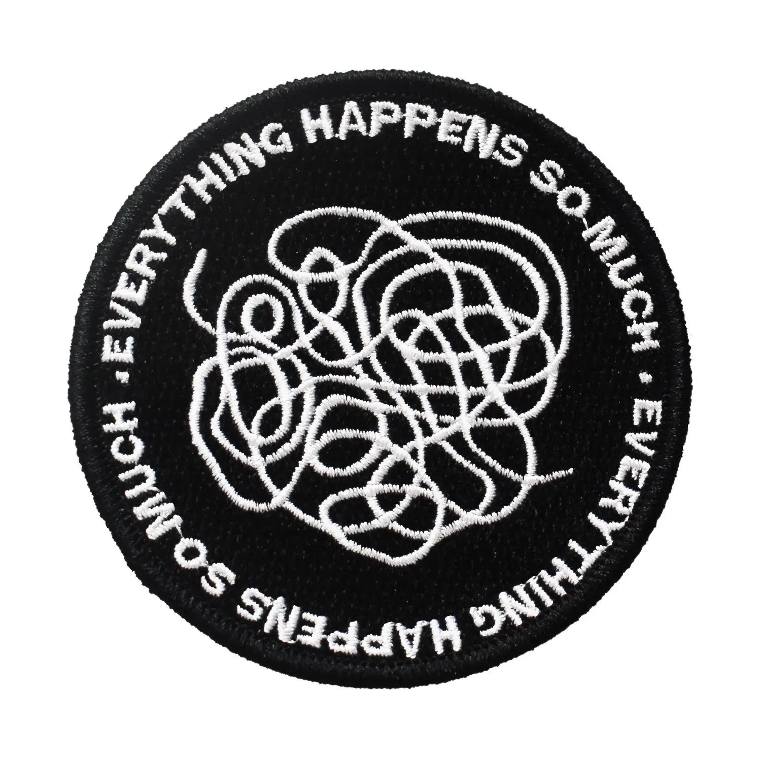 A round embroidered patch with a black background and the words “Everything happens so much” repeating around the border of the patch. There is an abstract swirled shape in white in the middle