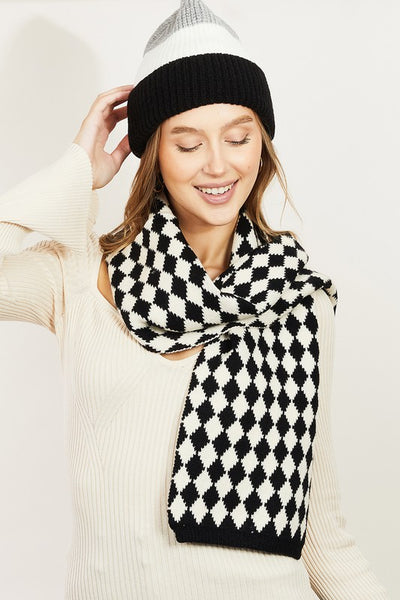 A knit black and creamy white scarf in a Harlequin pattern. Being worn wrapped around a model’s neck