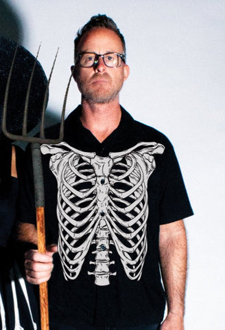 A black button down shirt with short sleeves and an ivory colored illustration on the front of a skeleton's rib cage. Shirt pictured on model