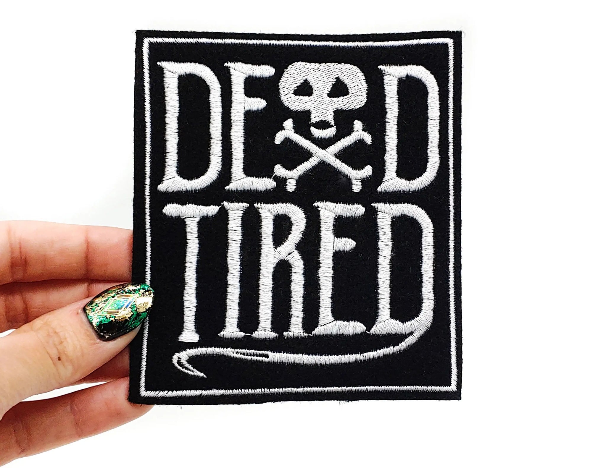 A rectangular embroidered patch with the words “DEAD TIRED” written in white stitching. The A in the word “dead” is a skull and crossbones 