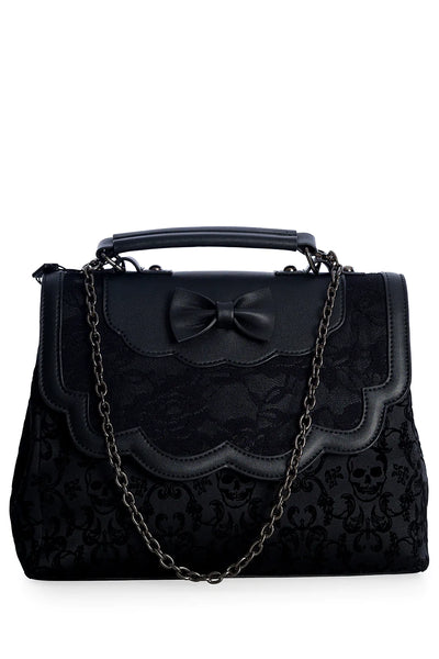 A black matte vinyl handbag with damask skull pattern body and a scalloped front flap with a bow and black lace detailing. The bag has a black matte vinyl hand strap and a gunmetal chain shoulder strap. The front of the purse with the shoulder strap hanging in front of the bag