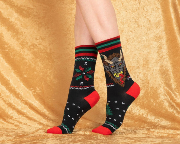 Fair Isle style pattern in red and green of holly and Christmas trees with an illustration of Krampus on soft black stretch cotton blend crew socks. Shows model wearing socks against a gold velvet background