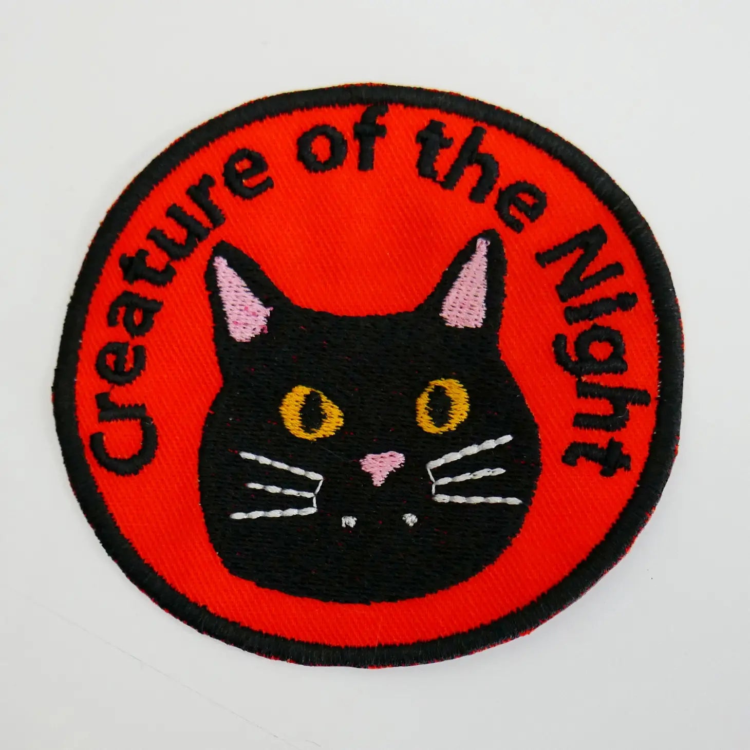 A round red twill patch with a black border and a black cat’s head embroidered in the middle with the message “Creature of the Night” embroidered in black above 