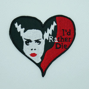 An embroidered patch portraying the Bride of Frankenstein with the lettering “I’d rather die” on a heart shaped patch with red background