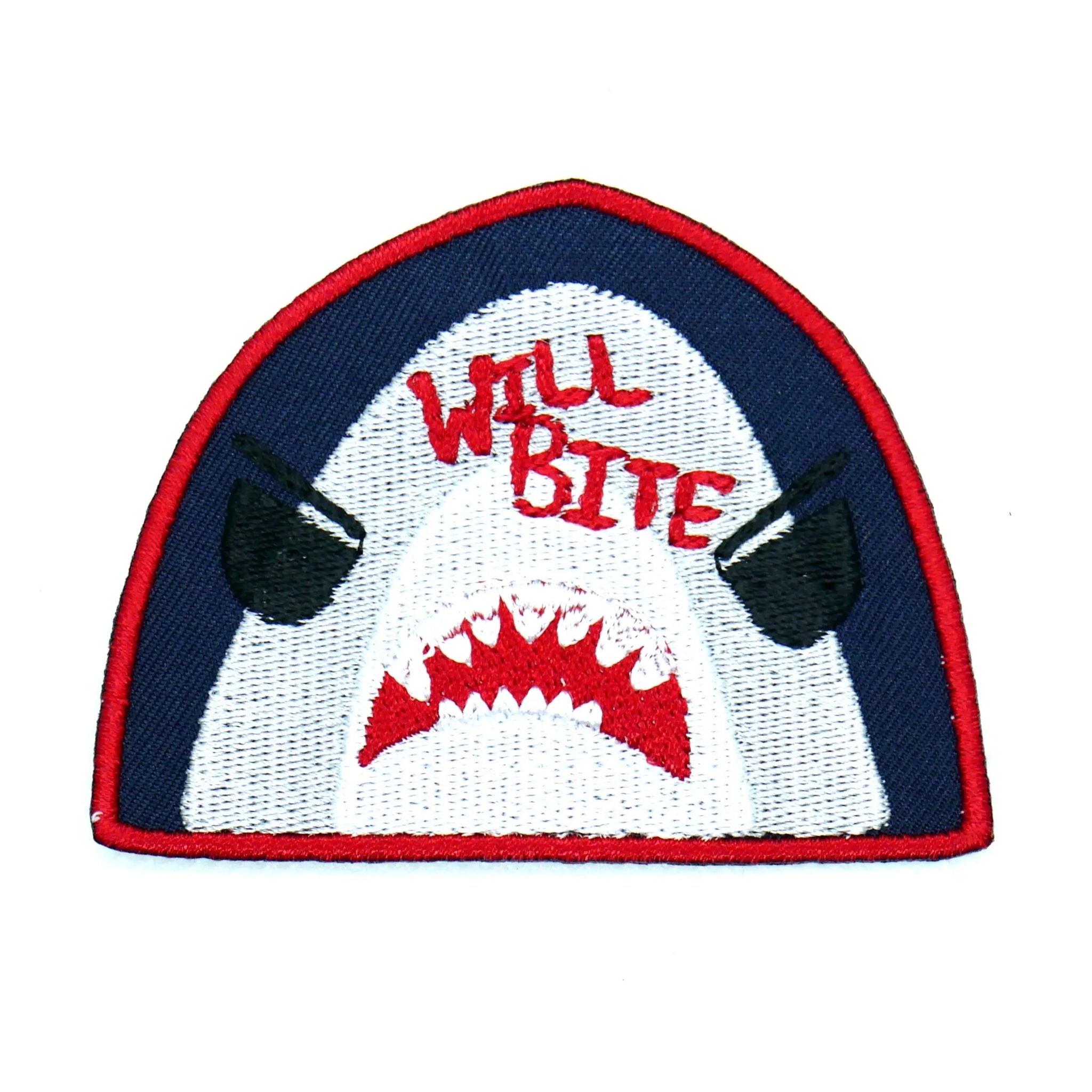 A navy blue cotton twill patch with a red border of a Great White shark with cartoon eyes and an open mouth. The words “Will Bite” are written in red at the top of the shark
