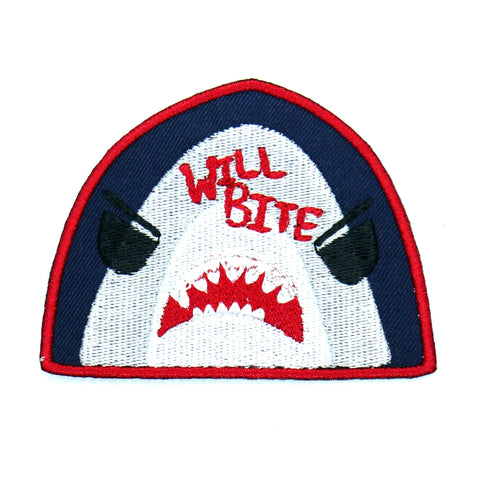 A navy blue cotton twill patch with a red border of a Great White shark with cartoon eyes and an open mouth. The words “Will Bite” are written in red at the top of the shark