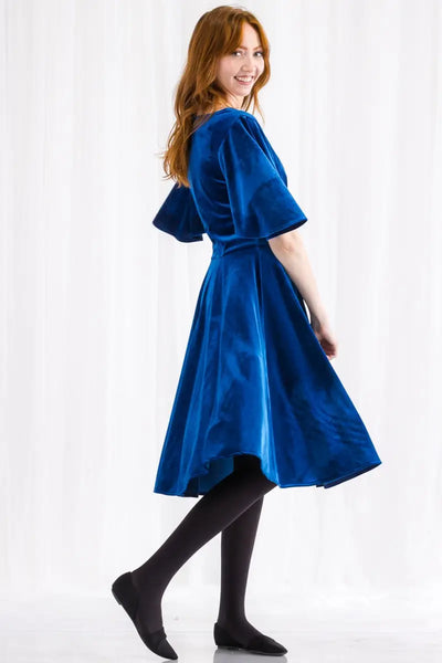 A royal blue velvet fit and flare dress with a deep plunging v sweetheart neckline, a wide banded waist, and elbow length flutter sleeves. Its skirt ends just past the knee. Shown on a model from a side angle