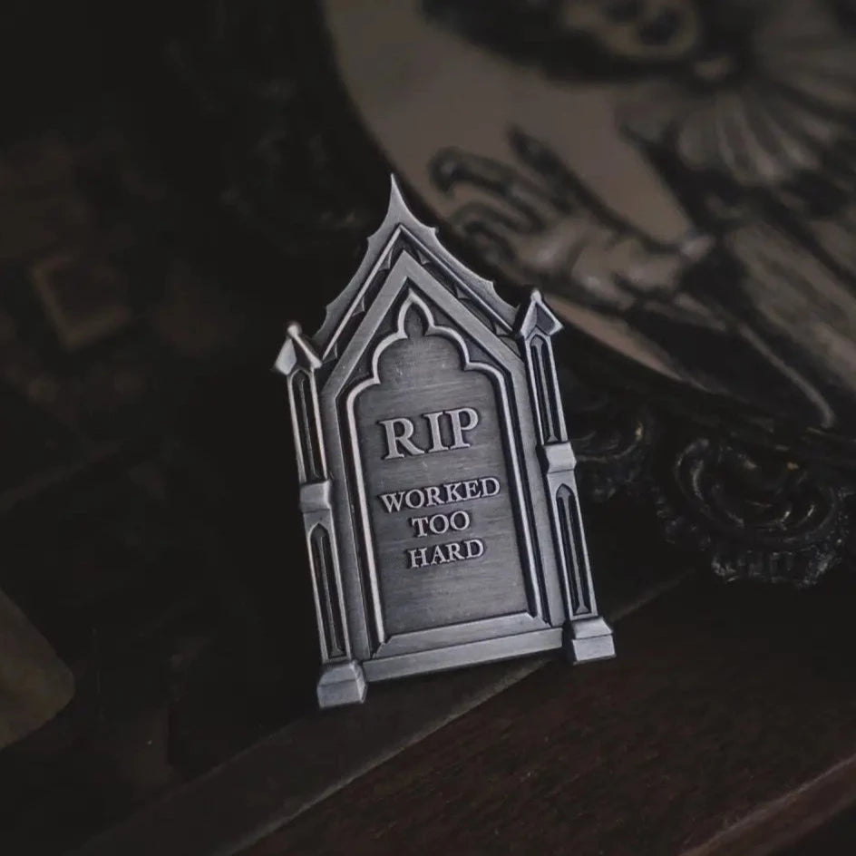 Antiqued silver finish metal pin in the shape of a gravestone with the epitaph of “RIP Worked Too Hard”