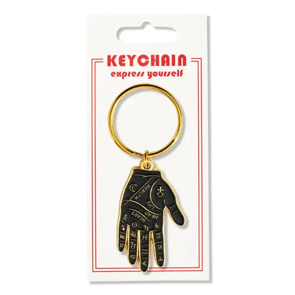 A shiny black and gold soft enameled hand pendant decorated with palmistry symbols on a sturdy gold metal keyring. Shown on its cardboard packaging
