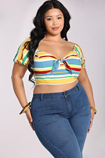 A plus size model wearing a short sleeved crop top with horizontal stripes in white, red, light blue, yellow, and seafoam green. It has a fitted bodice with tie detail and elasticized sweetheart neckline. 