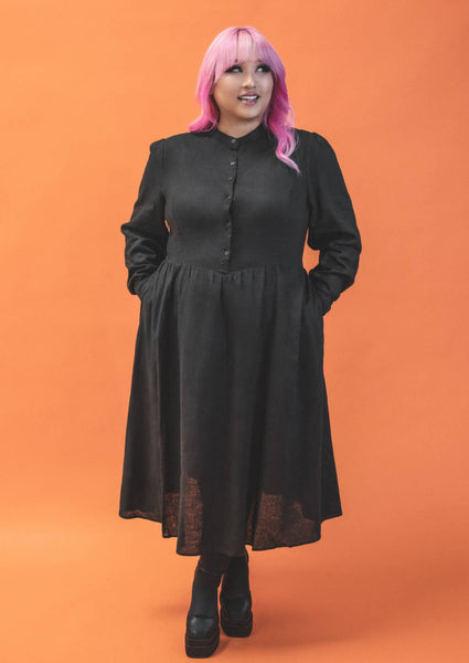 A plus size model wearing a long sleeved tea length linen shirtwaist dress with slightly puffed shoulders and princess seaming on the bodice. It has small matte black buttons down the front. The full skirt is slightly gathered. The model has their hands in both pockets