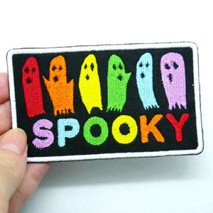 Rectangular patch with white border on a black background showing a row of ghosts in rainbow colors above the word “SPOOKY” also in rainbow lettering. Held by a hand