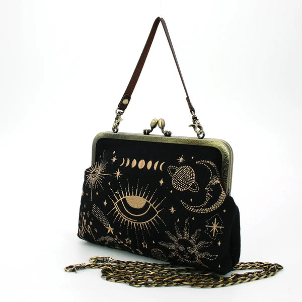 A vintage inspired handbag with an antiqued bronze metal kiss lock enclosure. The body of the purse is black fabric with gold embroidered planets, stars, moon phases, and hands. The handle is a detachable faux leather strap and in front of the bag is the antiqued bronze metal shoulder strap. Shown from the side to illustrate width of the purse