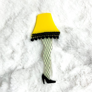 acrylic brooch of the iconic leg lamp from Bob Clark’s 1983 movie A Christmas Story with intricate hand-painted fishnet detail and pom-pom trim along the edge of the bright yellow lampshade