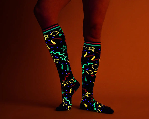A pair of black knee socks with neon blue, green, and pink stripes at the cuff and blue toes & heels. There is a neon colored geometric pattern along the calf of each sock similar to an 80s bowling alley carpet. Shown on a model in UV lighting 
