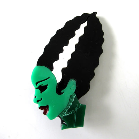 A layered lasercut acrylic brooch of the side profile of the Bride of Frankenstein in black, white, red, and various shades of green