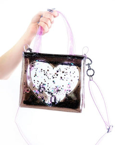 A hand holding a vinyl purse with a black glitter vinyl body and a large clear heart with a mix of multicolored glitter encased in liquid. The purse has pink vinyl hand and cross body straps