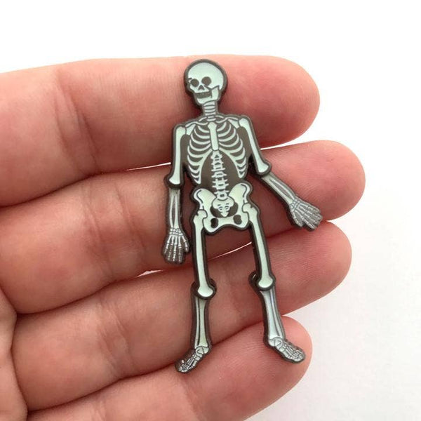 shiny gunmetal and glow-in-the-dark white enamel skeleton lapel pin with moving elbow and knee joints, shown held in a hand