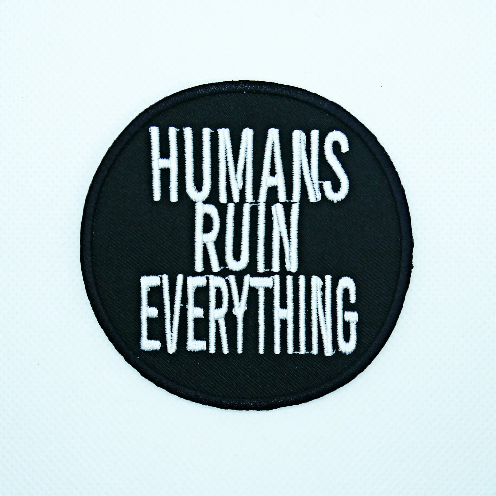 Round embroidered patch with the phrase “HUMANS RUIN EVERYTHING” in white capital letters on a black background