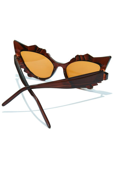 A pair of cat-eye sunglasses sitting at an angle that displays their arms. They are a translucent warm brown color with geometric detail above and below the lenses, which are brown 
