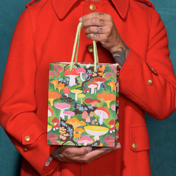 A gift bag with an allover print of beautiful multicolored mushrooms in a scenic nature background, illustrated by Kelsey Garrity Riley. held by a model to show size.
