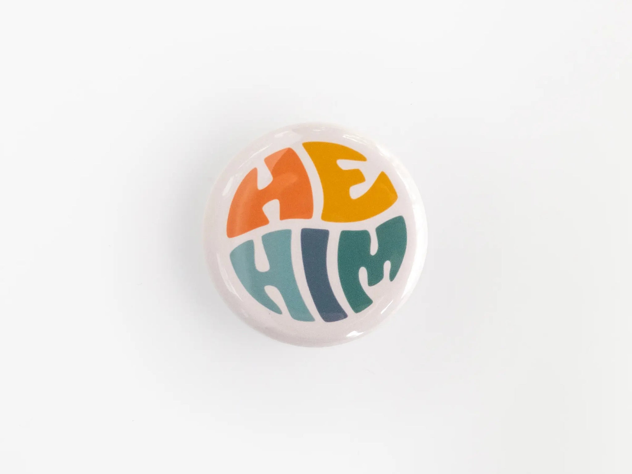 A 1.25” button with the pronouns “he” and “him” in multicolored lettering