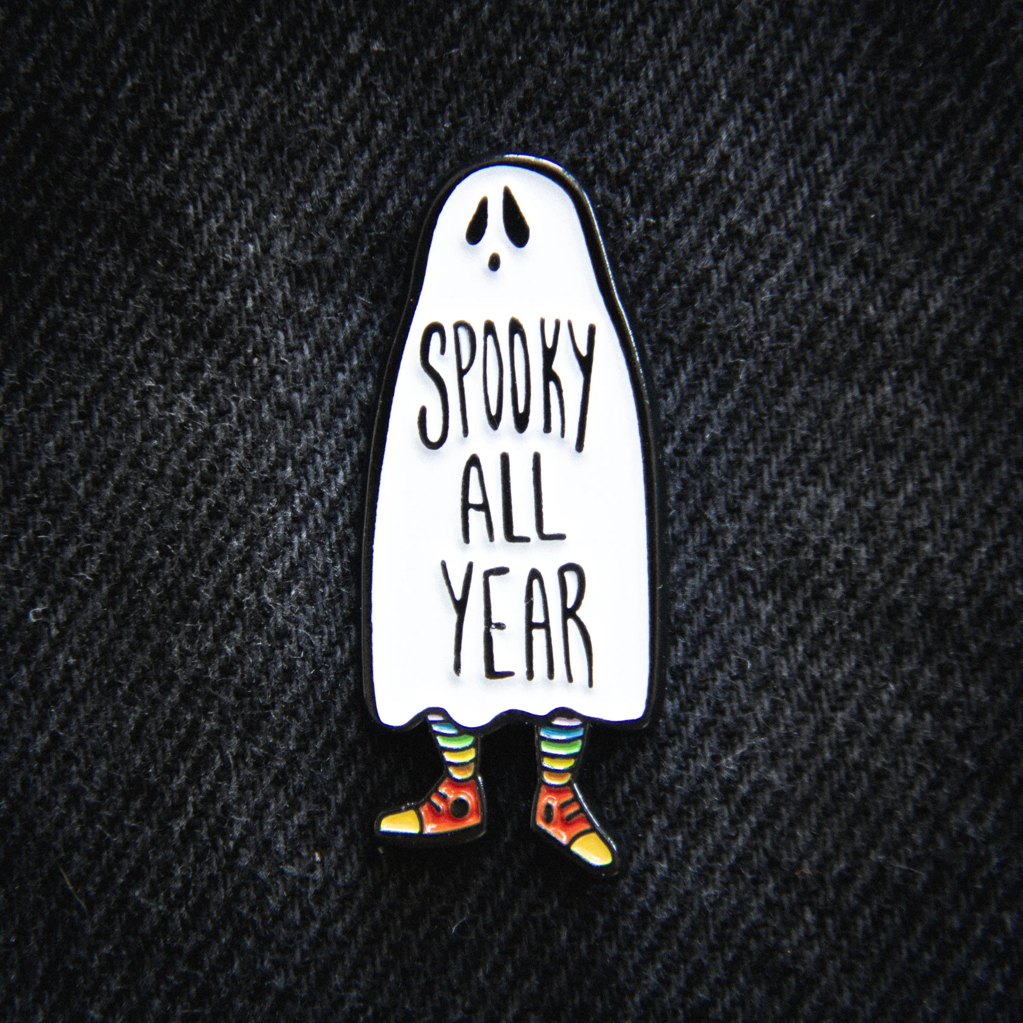 An enamel pin of a ghost wearing rainbow socks and red high top sneakers. The words “SPOOKY ALL YEAR” are written across its middle