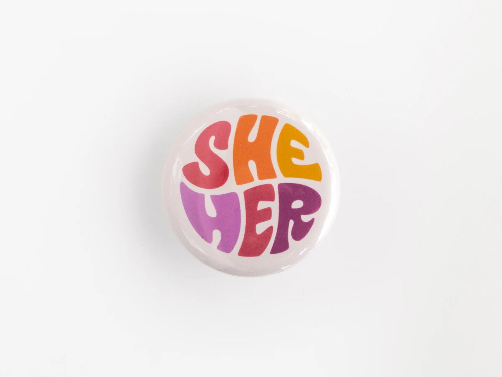 A 1.25” button with the pronouns “she” and “her” in multicolored lettering