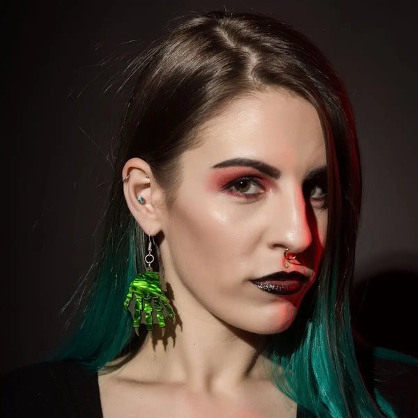 A pair of laser-cut hand layered acrylic dangle earrings In the shape of the Creature from the Black Lagoon’s claws. With marbled green with matte green webbed claws & engraved and hand painted gold detailing. Shown on a model 