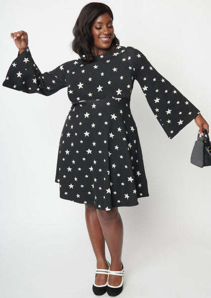 A black sweater knit fit and flare dress with raglan bell sleeves in a black and white star pattern. Shown on a model with their arms raised to show bell sleeves