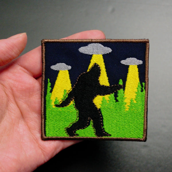embroidered patch of Sasquatch walking silhouette against background of green forest trees and three UFOs in the night sky, held in a hand