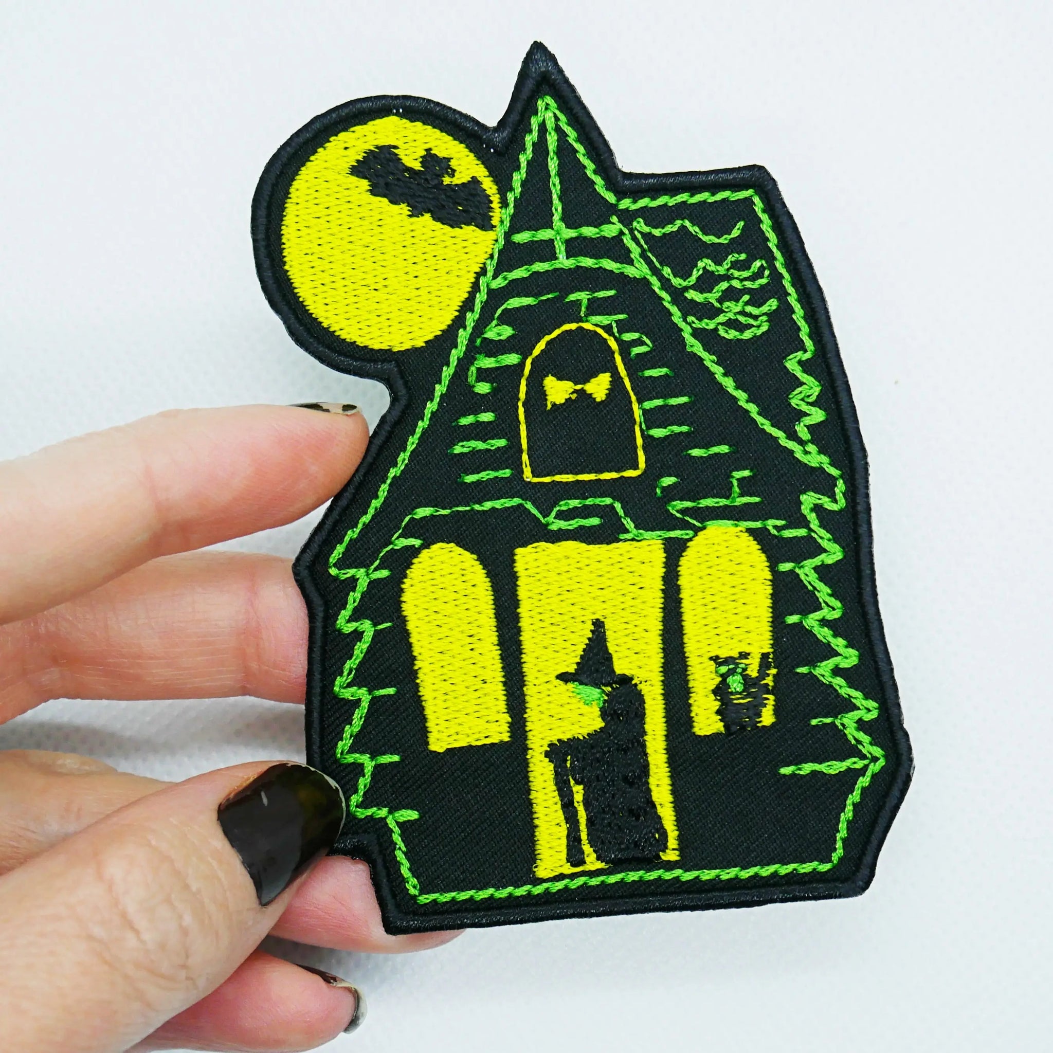 A hand holding a black canvas twill patch of a green and yellow haunted house with a witch standing in the doorway & a yellow full moon rising in the background 