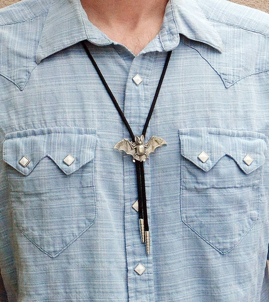 silver metal bat-shaped bolo tie on black cord, shown worn with light blue denim shirt on model