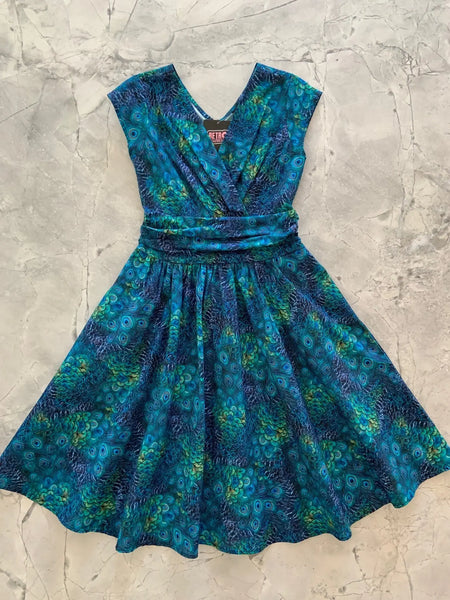 A cap sleeve cotton dress in a green and blue peacock feather print with a surplus gathered neckline, wide gathered waist, and a just past the knee full skirt. Seen laid flat 