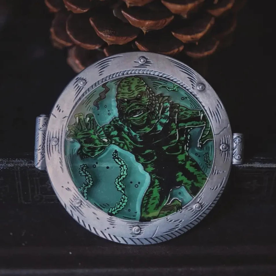Round enamel pin in the shape of a silver porthole on a boat with the Creature from the Black Lagoon swimming in a background of translucent blue water.