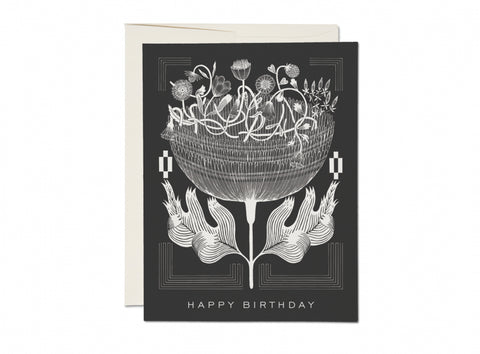 Greeting card with a black and white illustration of a large poppy with smaller poppies and clover at the top of the blooming flower. Surrounded by white geometric designs on a black background with the lettering “Happy Birthday” at the bottom of the card