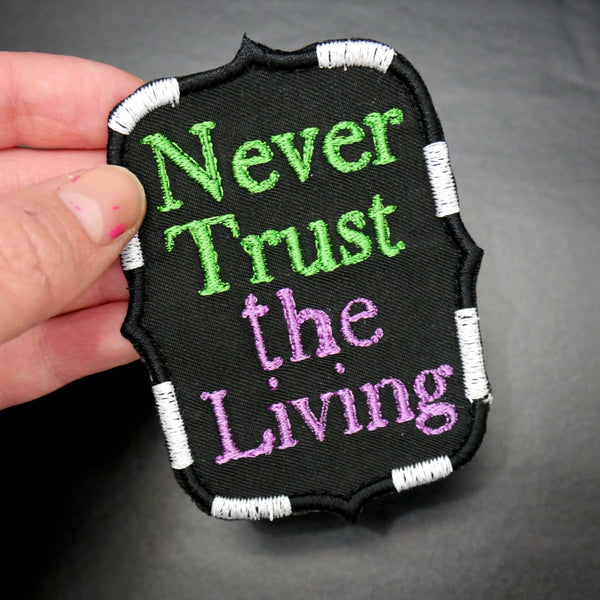 black canvas patch with "Never Trust the Living" text in green and purple with black and white stripe border, held in hand to show size
