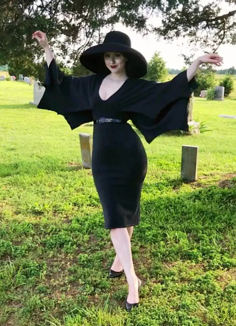 A model wearing a black wiggle length dress with a rounded v neck, scalloped sleeves that resemble a bat’s wings, and a black patent belt. They are standing in a cemetery with their arms raised posing