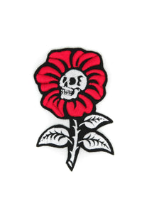An embroidered patch of a flower with a thorny stem and bright red petals. In the middle of the flower there is a white skull