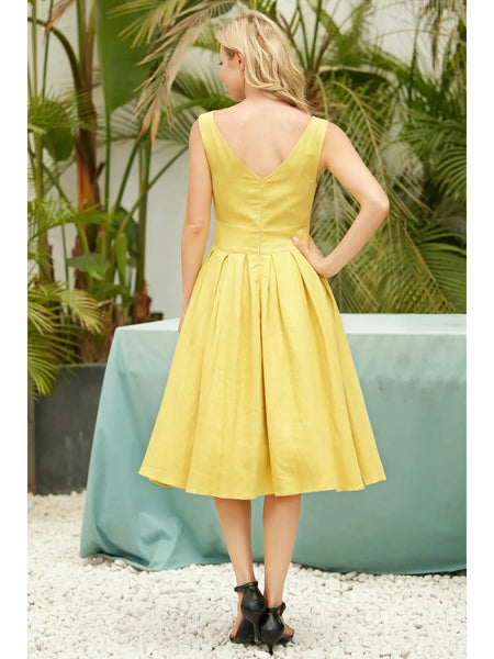 A model wearing a sleeveless dress in a shade of golden yellow with a fitted high neckline and princess seamed bodice, wide banded waist, full box-pleated just below the knee length skirt. Seen from behind