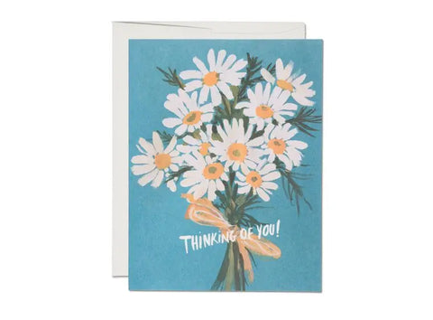 Greeting card of an illustration of a bouquet of daisies tied with a yellow ribbon on a blue background. Lettering of “Thinking Of You!” at the bottom of the card
