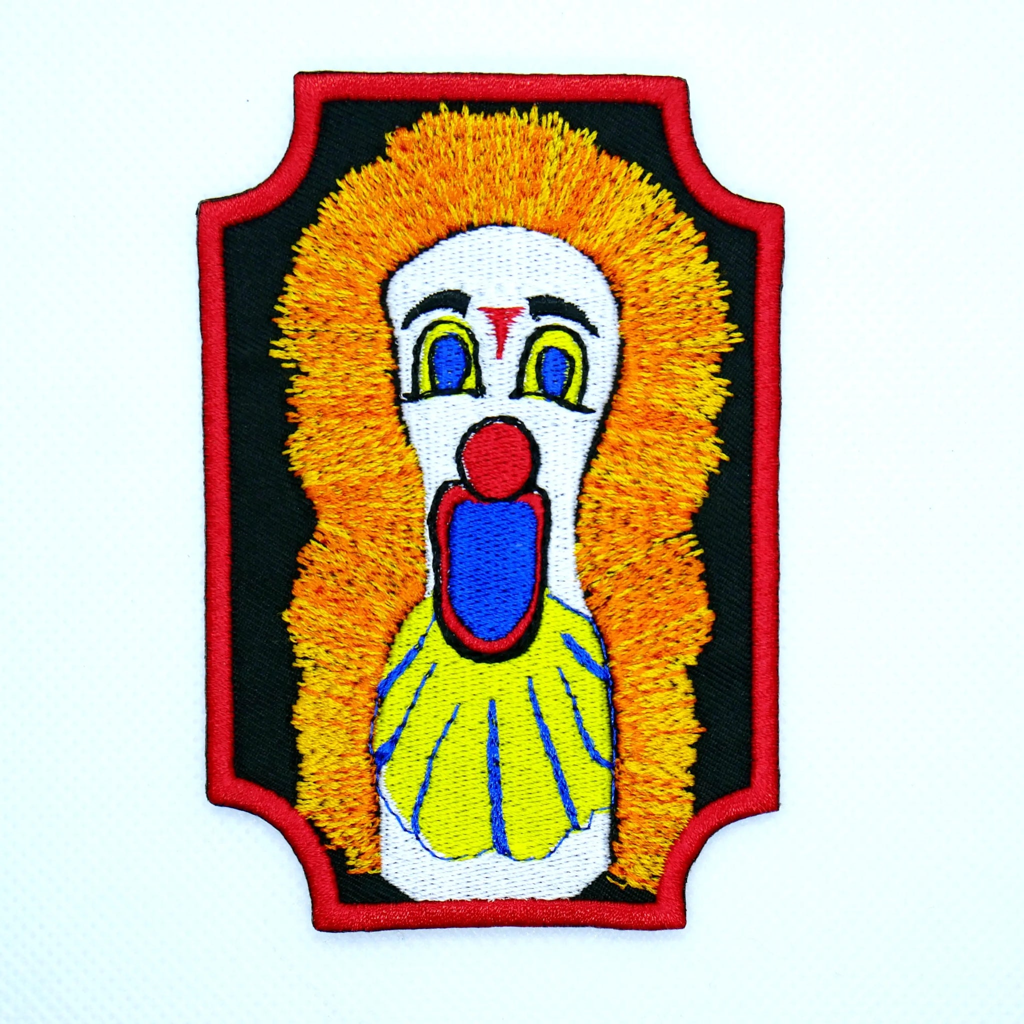 A rectangular black twill embroidered patch with scalloped edges and a bright red border featuring a white, blue, and red carnival style clown with an open mouth and bright orange hair.