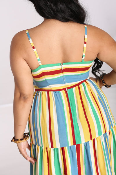 A plus size model wearing a sleeveless sundress with horizontal red, blue, yellow, white, and seafoam stripes. It has adjustable spaghetti straps, a sweetheart neckline, bamboo ring detail at the bodice, and a three tiered long skirt. Shown from behind to feature zip back closure
