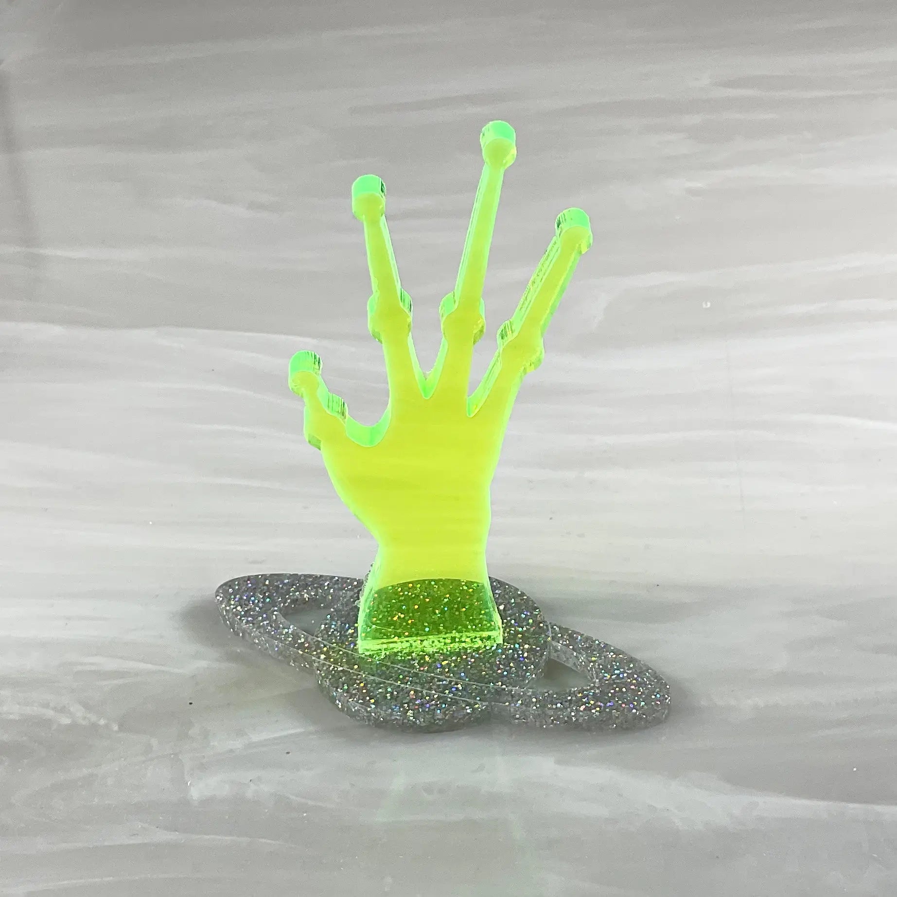 A ring holder made of acrylic in the shape of a neon green alien hand on a silver glittery Saturn shaped base.